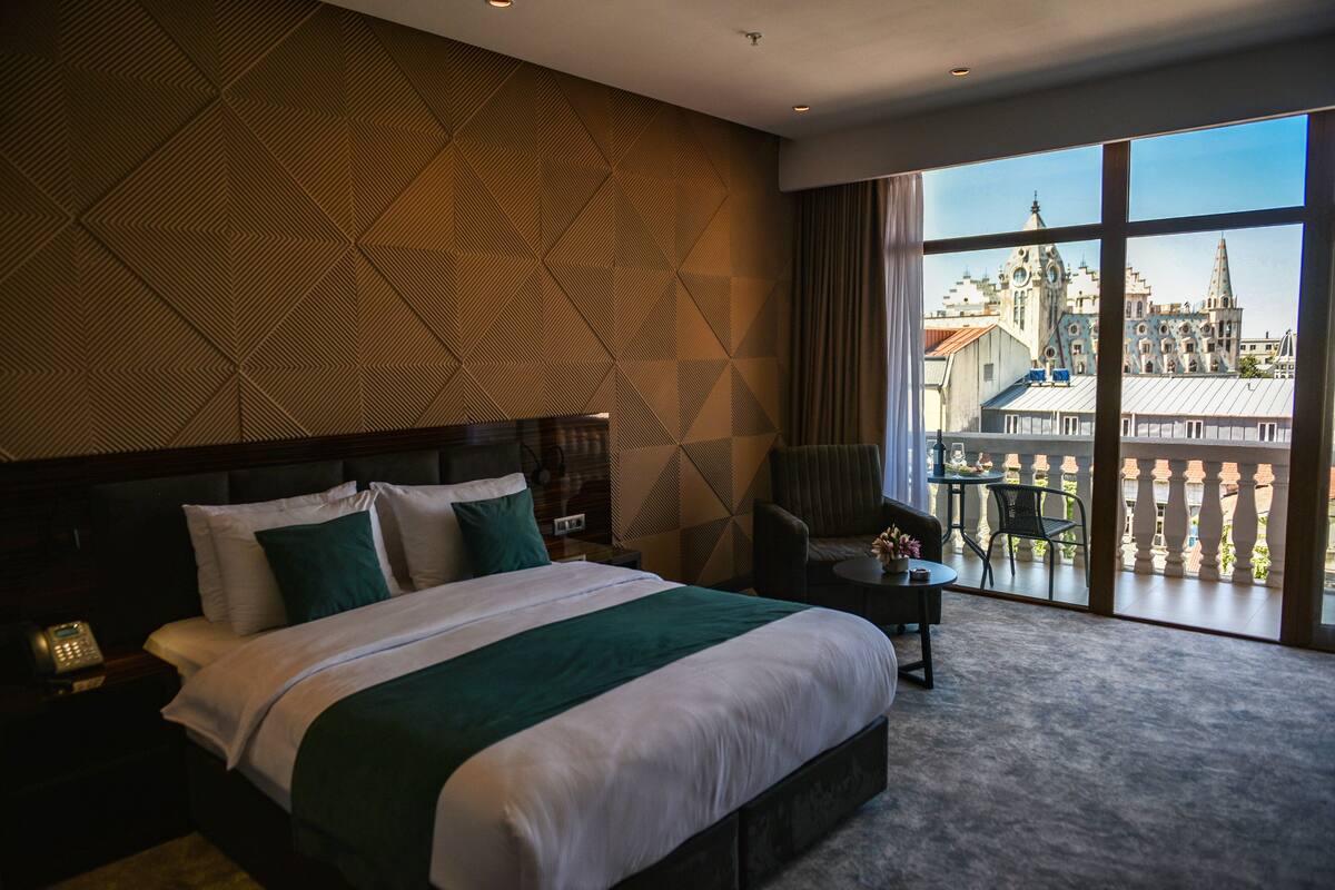 Habitación de hotel con una cama grande, una mesita de noche, una silla y vistas de un paisaje urbano a través de amplias ventanas.