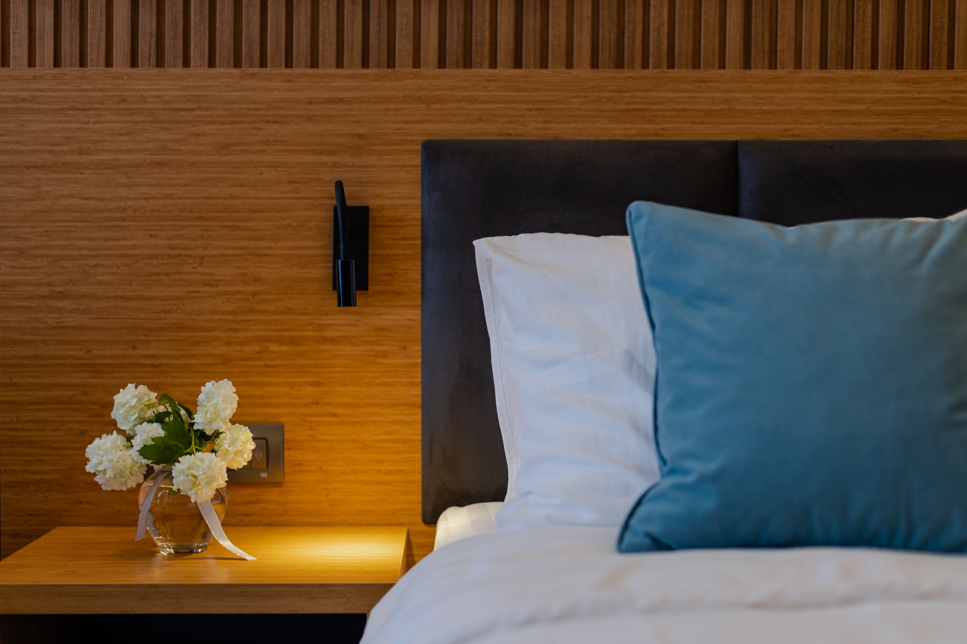 A neatly made bed with white and blue pillows, a small vase with white flowers on a wooden nightstand, and a wall-mounted light.