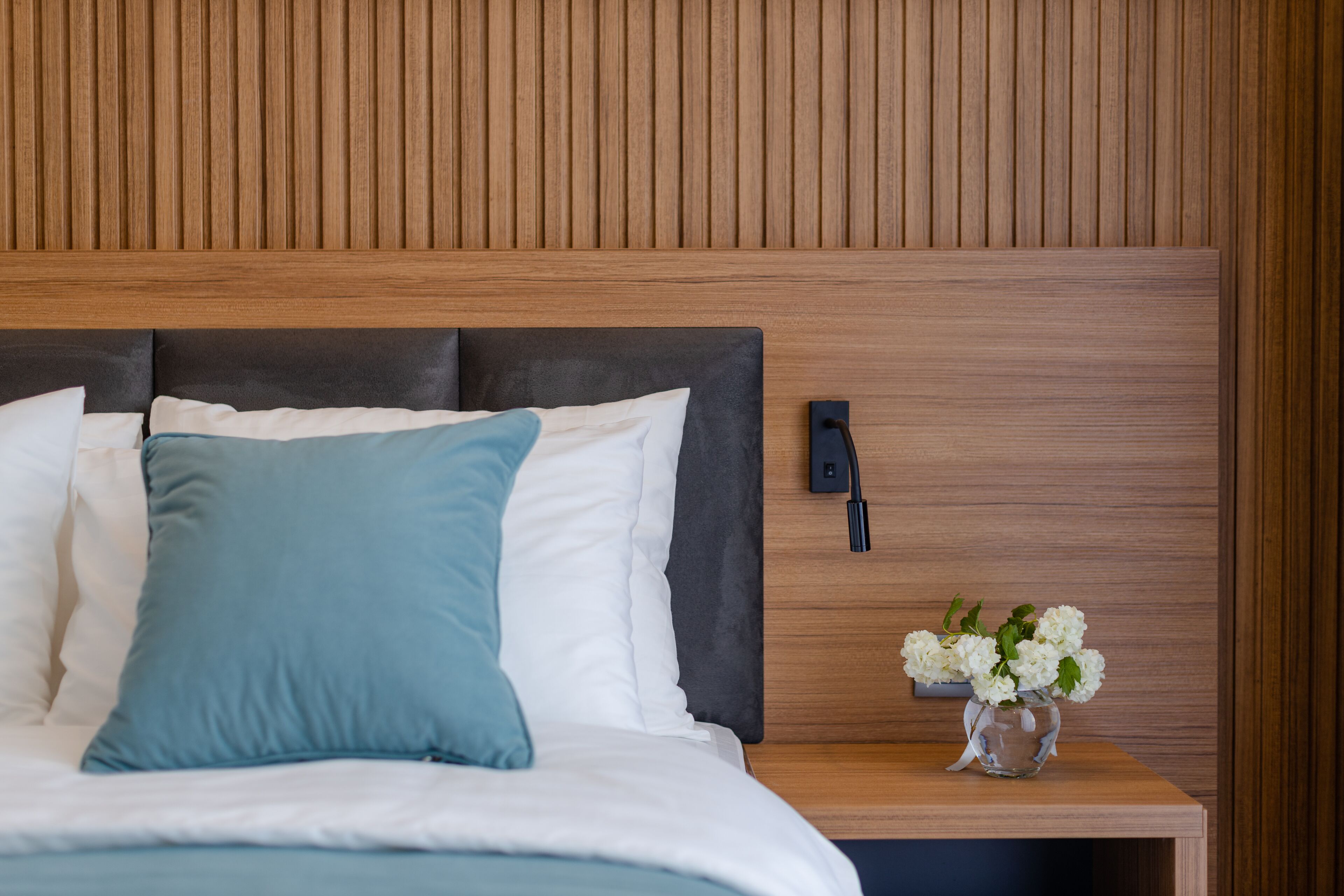 A neatly made bed with white and blue pillows, a wooden headboard, and a nightstand with a vase of white flowers.