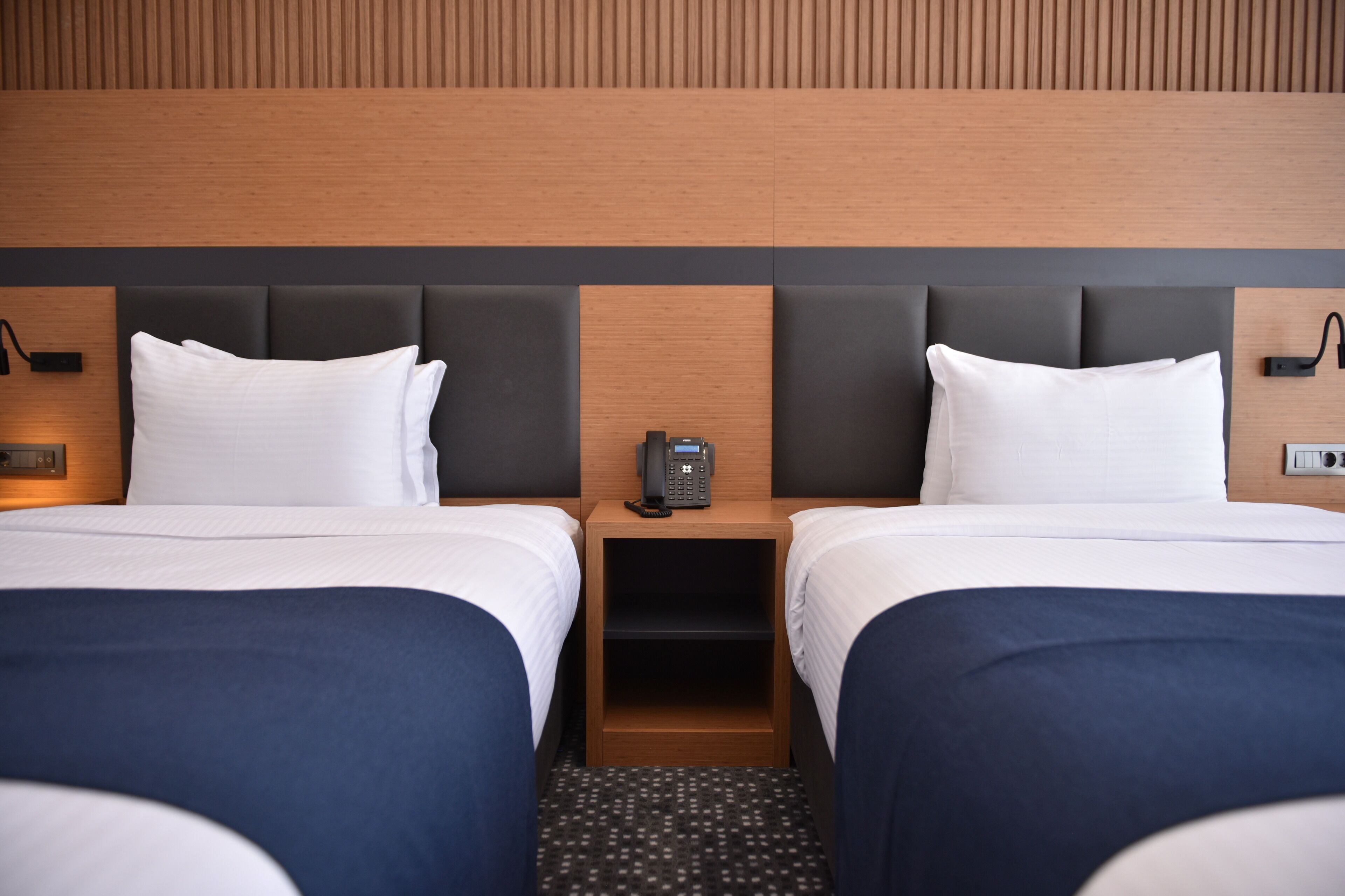 Ein Hotelzimmer mit zwei Betten, einem hölzernen Kopfteil, einem Nachttisch mit Telefon und einem gemusterten Teppich.
