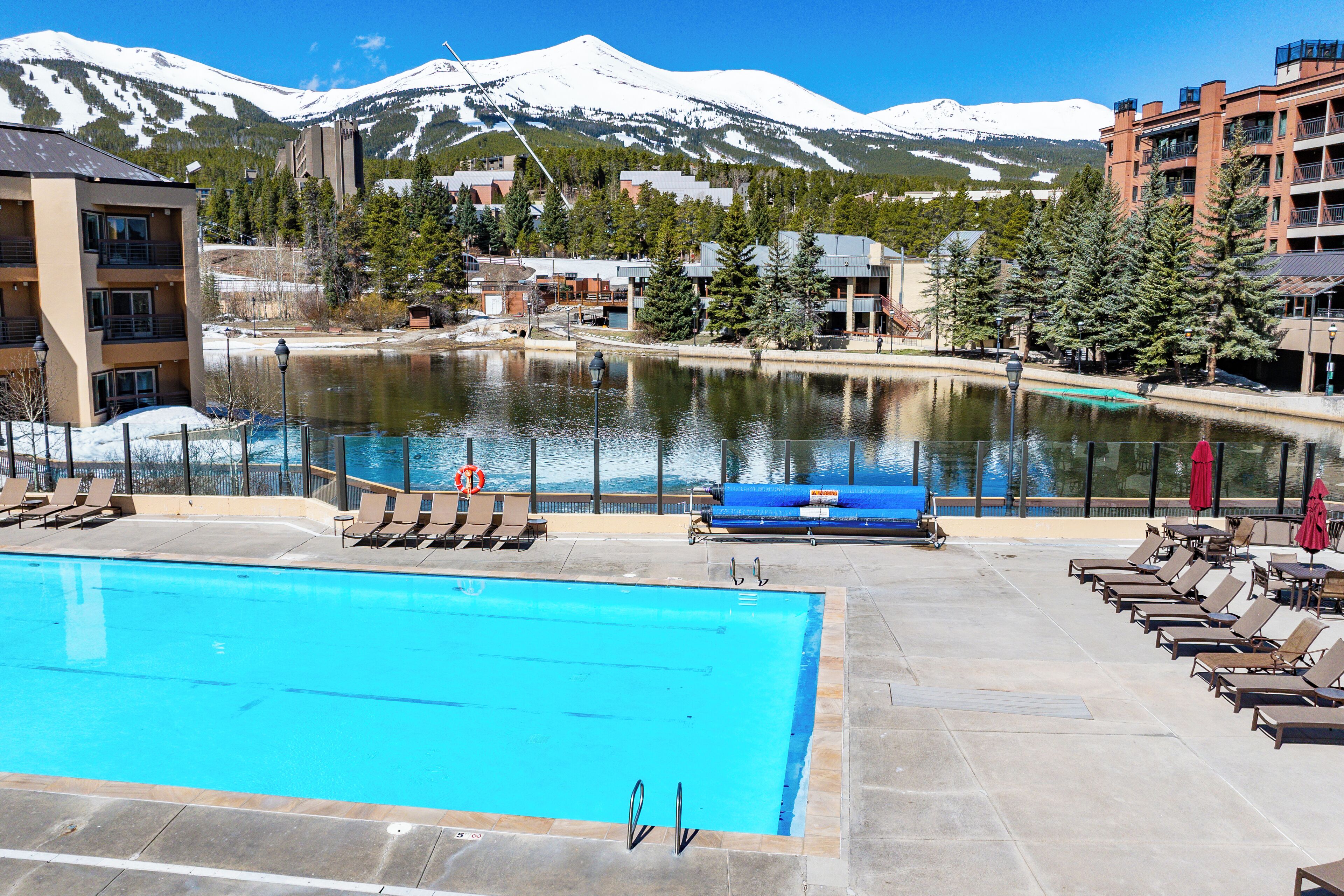 A swimming pool with lounge chairs and umbrellas, a view of a lake, and buildings in the background with snow-capped mountains.
