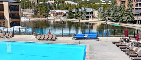 A swimming pool with lounge chairs and umbrellas, a view of a lake, and buildings in the background with snow-capped mountains.