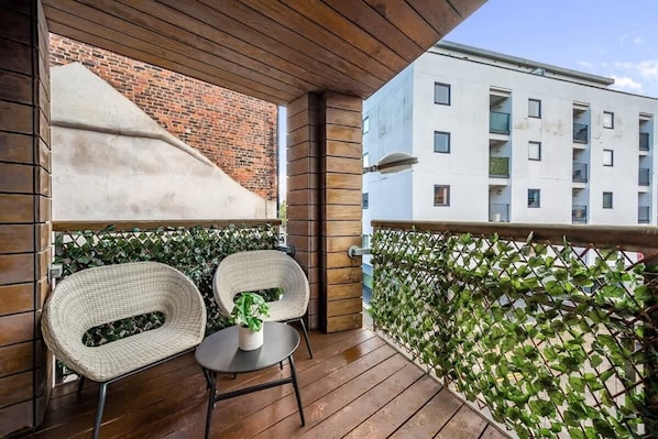 A balcony with two wicker chairs, a small table, and a potted plant. A brick wall and a white building with balconies are visible in the background.