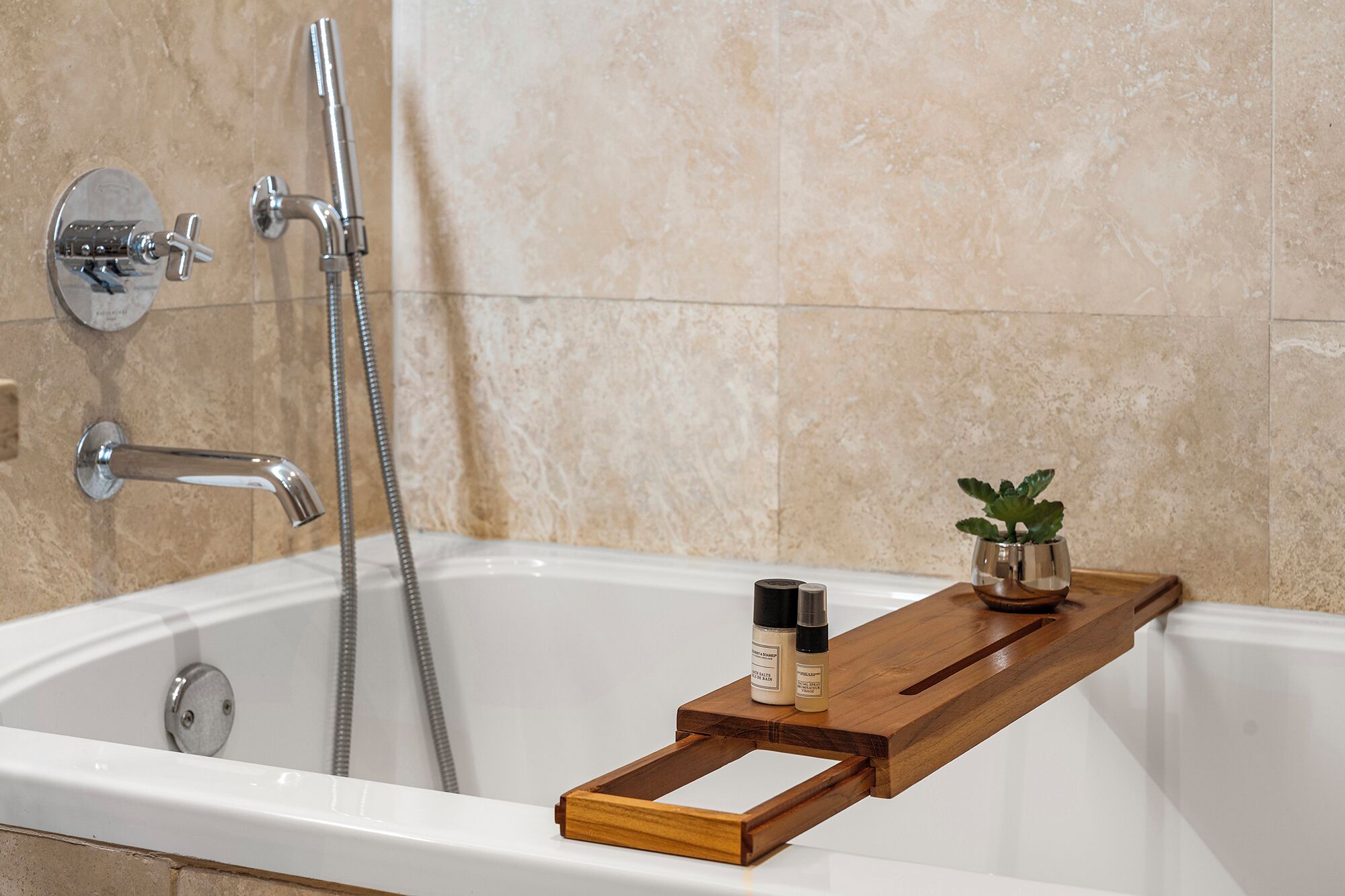 Un baño con bañera, ducha de mano, estante de madera con artículos de aseo y una pequeña planta en maceta.