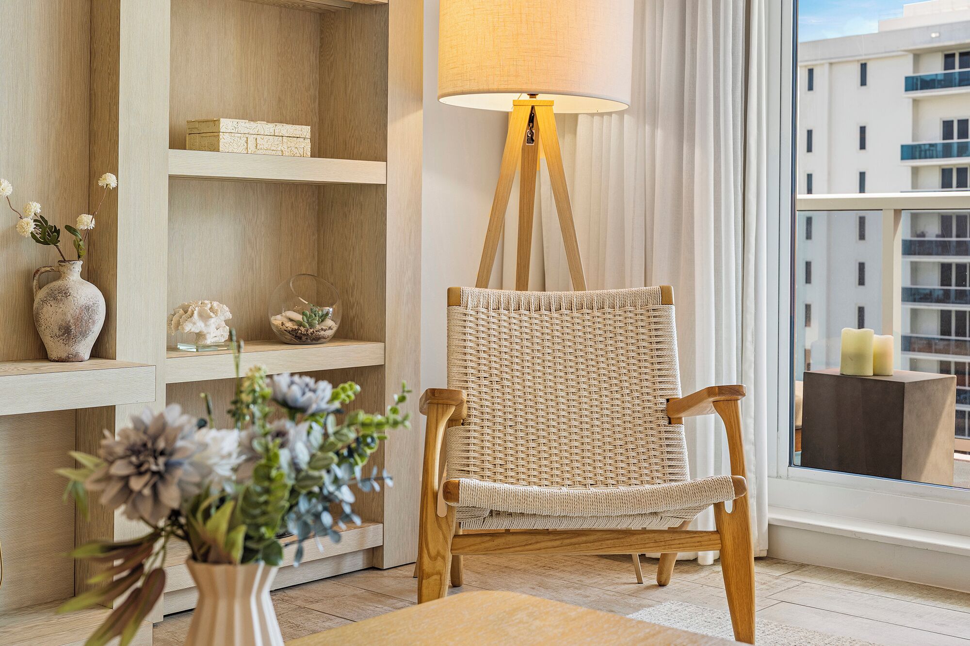 Une pièce avec un fauteuil en osier, des étagères en bois, un vase rempli de fleurs et une lampe.