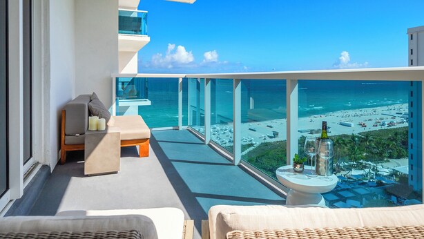 A balcony with a view of the beach, a sofa, a small table with a bottle and glasses, and a chair.