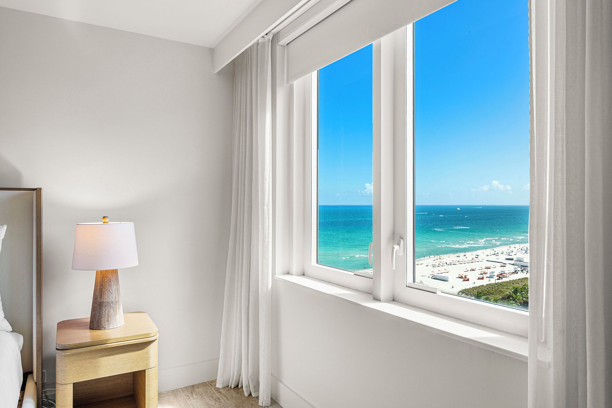 Ein Zimmer mit großem Fenster und Blick auf einen Strand, einem Nachttisch mit Lampe und einem Bett.