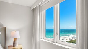 A room with a large window overlooking a beach, a bedside table with a lamp, and a bed.