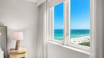Ein Zimmer mit großem Fenster und Blick auf einen Strand, einem Nachttisch mit Lampe und einem Bett.