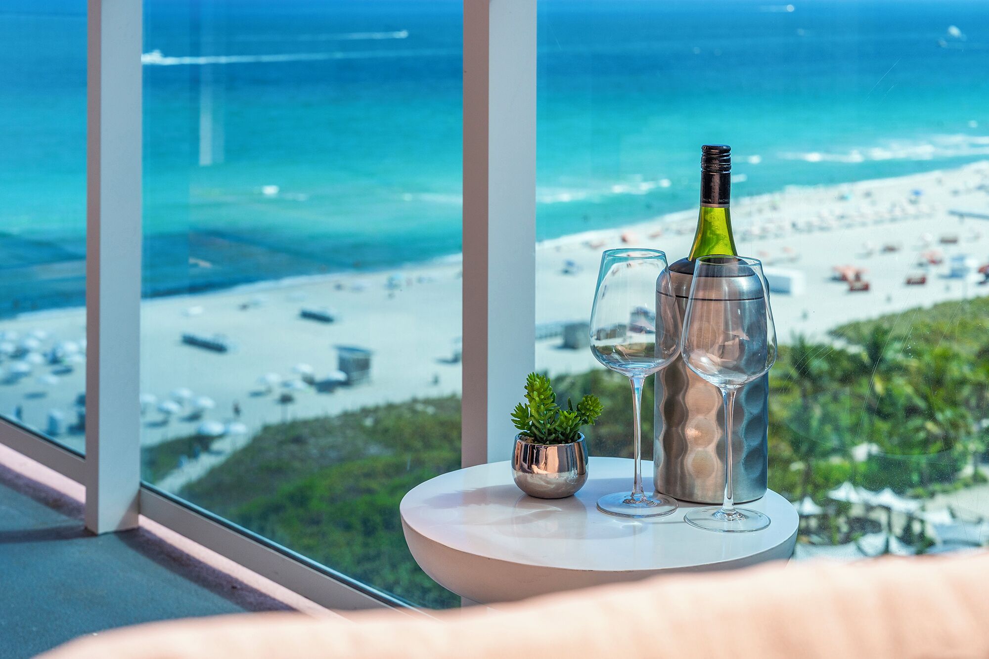 Una mesa con una botella y vasos, una planta pequeña, y vistas a una playa y al océano.