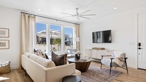 Un salon moderne avec un canapé, une table basse et un fauteuil. Un téléviseur est fixé au mur et un ventilateur de plafond est installé.