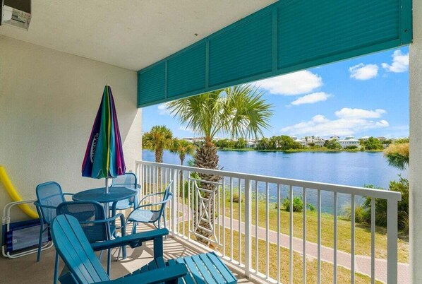 Un balcón con sillas azules y una mesa, con vista a un lago con palmeras y casas al fondo.