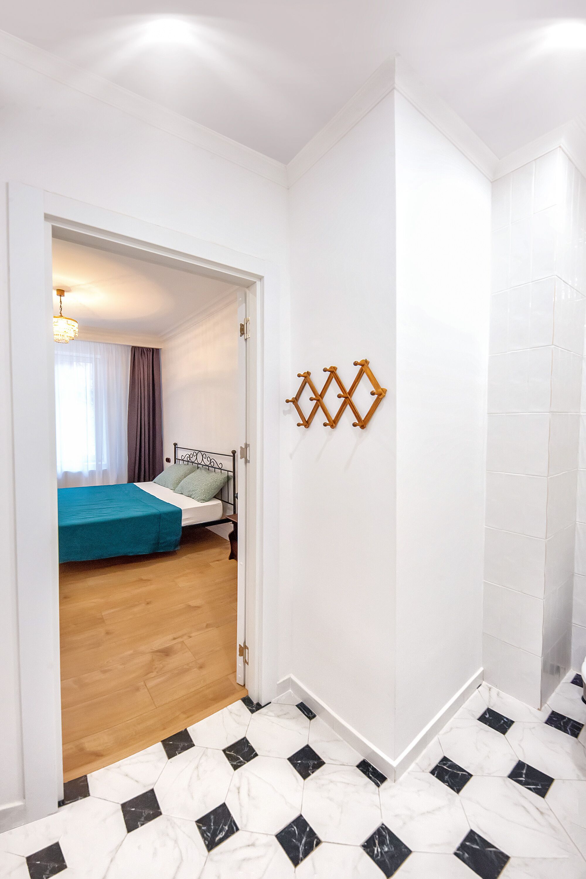 Habitación de hotel con piso de madera, cama con cubrecama azul, pared blanca con decoración geométrica y pasillo con baldosas de diseño blanco y negro.