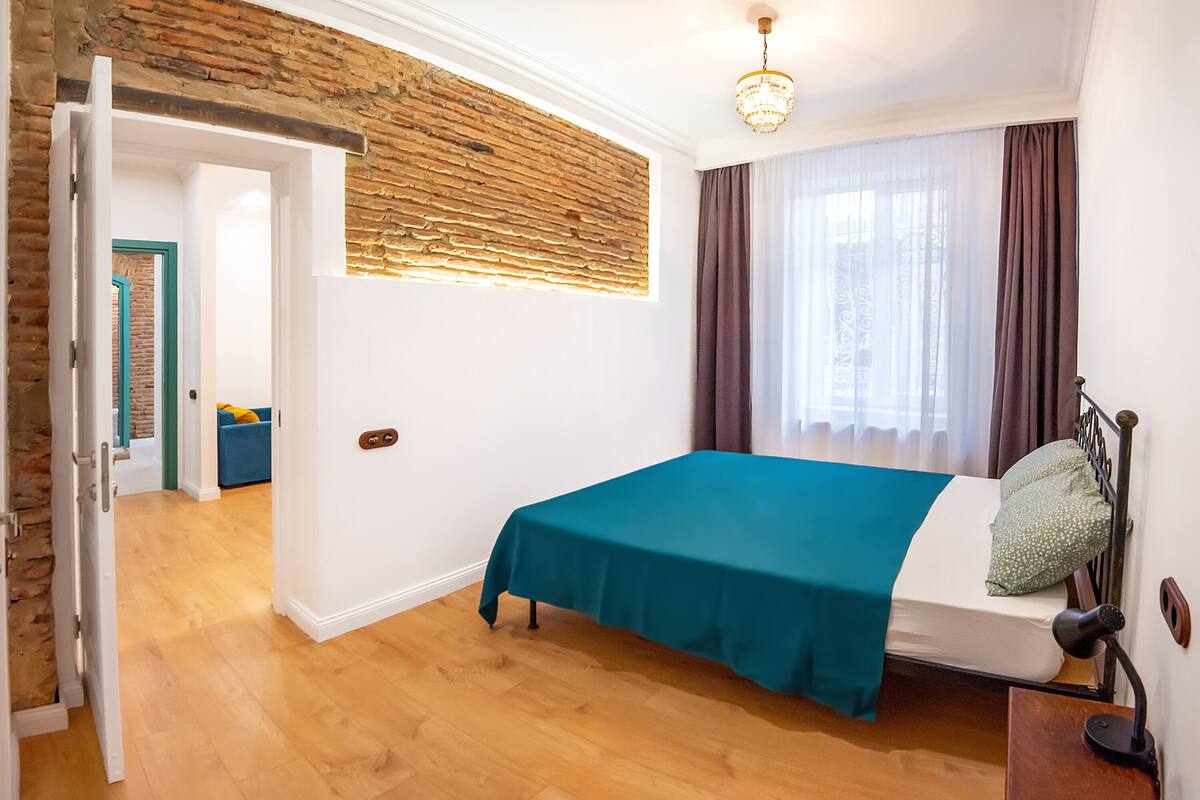 A bedroom with a bed, a wooden floor, a brick wall, a window with curtains, and a door leading to another room.
