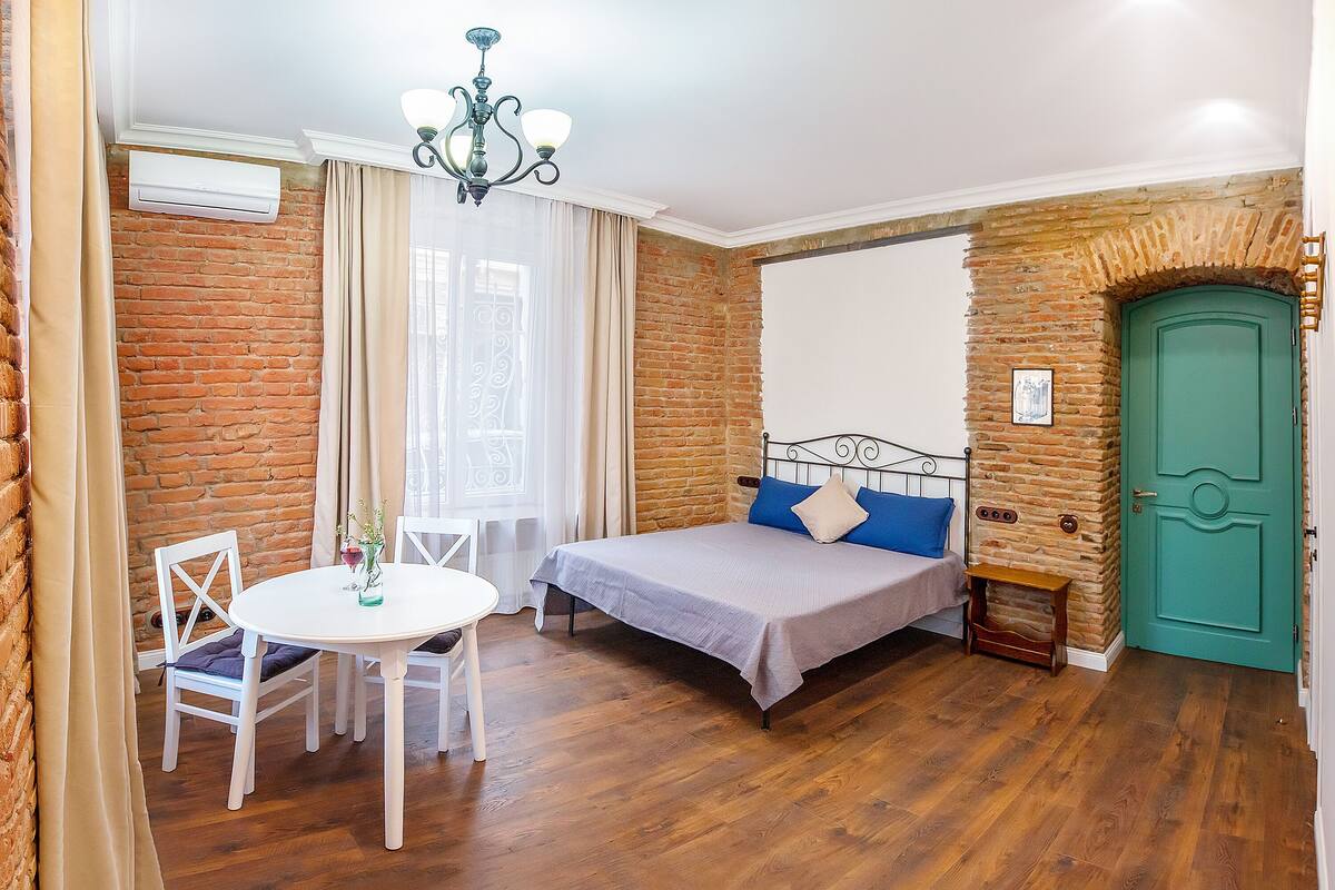 A room with a bed, a table, and a chair. There is a brick wall and a green door.