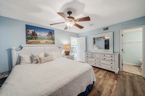 A bedroom with a bed, a ceiling fan, a dresser, a mirror, and a door leading to a bathroom.
