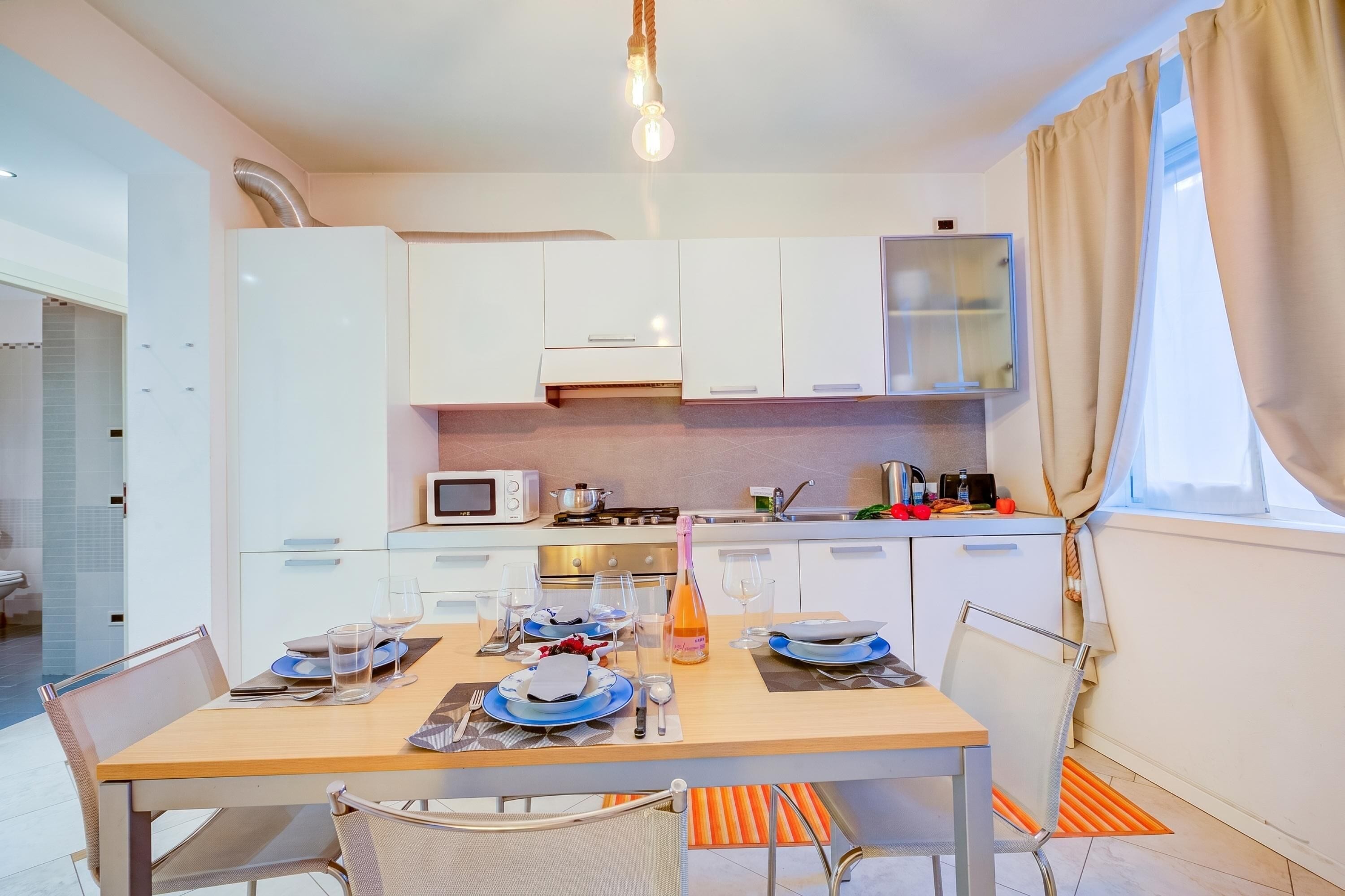 A modern kitchen with white cabinets, a dining table set for four, and a large window with curtains.