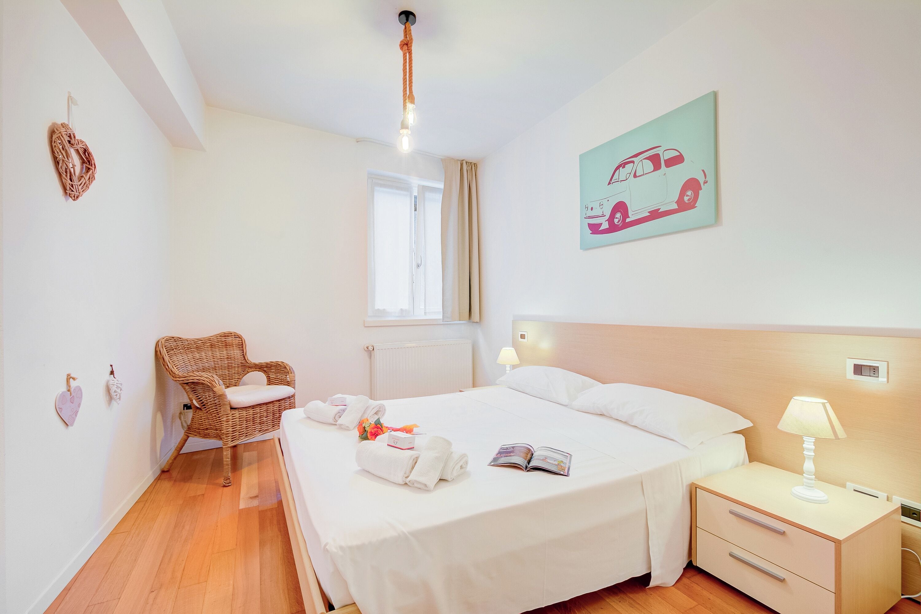 A hotel room with a bed, a nightstand, a wicker chair, a window, and a wall art of a car.