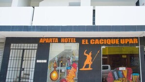 Fachada del Aparta Hotel El Cacique Upar, con un letrero destacado y una colorida exhibición en las ventanas que muestra instrumentos musicales.