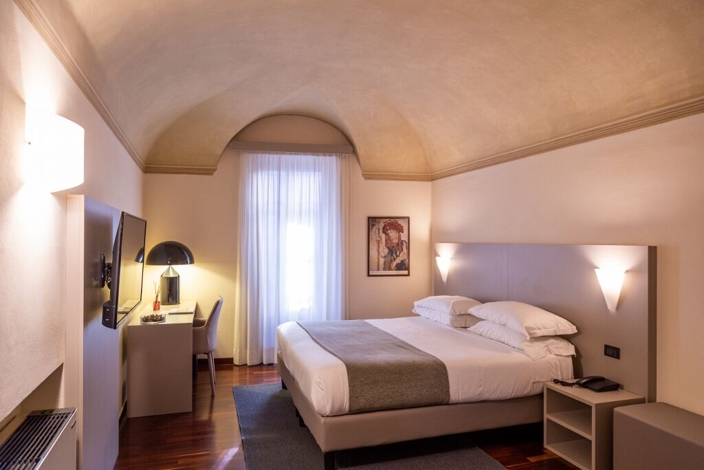 Una habitación de hotel con cama, mesita de noche, lámpara, escritorio, silla y un cuadro en la pared.
