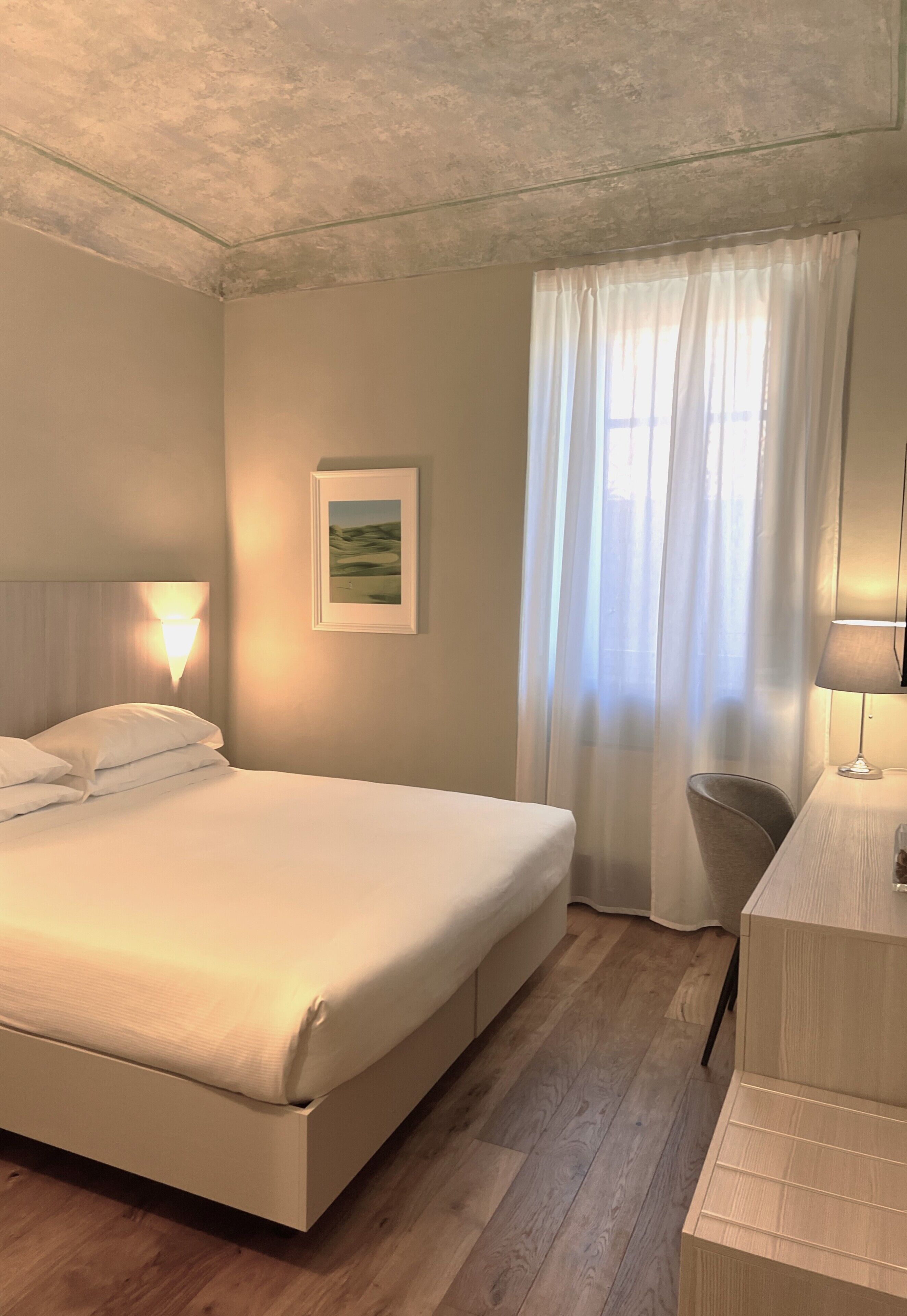 Habitación de hotel con una cama grande, una mesita de noche, una lámpara, un cuadro en la pared y una ventana con cortinas traslúcidas.