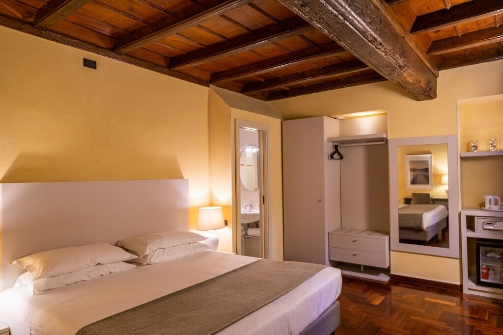 Una habitación de hotel con cama, lámparas de noche, un espejo, armario y una pequeña kitchenette.