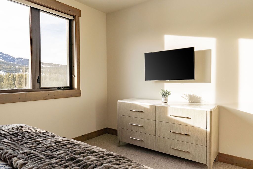 Un dormitorio con cama, cómoda, televisor de pantalla plana y ventana con vistas a árboles y montañas.