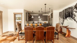 A dining area with a wooden table and chairs, a chandelier, and a large painting on the wall. - Luxury 4BR Big Sky Condo | Steps to Resort, Hot Tub & America's Best Skiing (Big Sky)