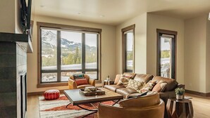 A spacious living room with a large window offering a mountain view, a leather sofa, a coffee table, and a fireplace. - Luxury 4BR Big Sky Condo | Steps to Resort, Hot Tub & America's Best Skiing (Big Sky)