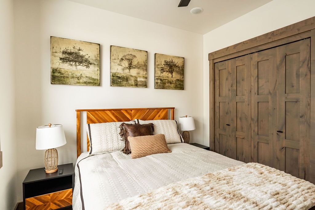 Un dormitorio con cabecera de madera, una cama grande con manta blanca y beige, dos mesitas de noche con lámparas y tres cuadros de árboles colgados en la pared.