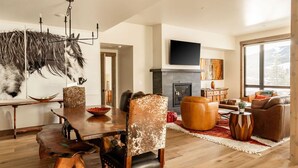 A spacious living room with a fireplace, a dining area with a wooden table and chairs, and a large window offering a view of the outdoors. - Luxury 4BR Big Sky Condo | Steps to Resort, Hot Tub & America's Best Skiing (Big Sky)