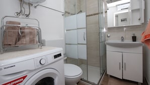 Ein modernes Badezimmer mit Waschmaschine, einer Duschkabine aus Glas, Toilette und Waschbecken mit Spiegel darüber.