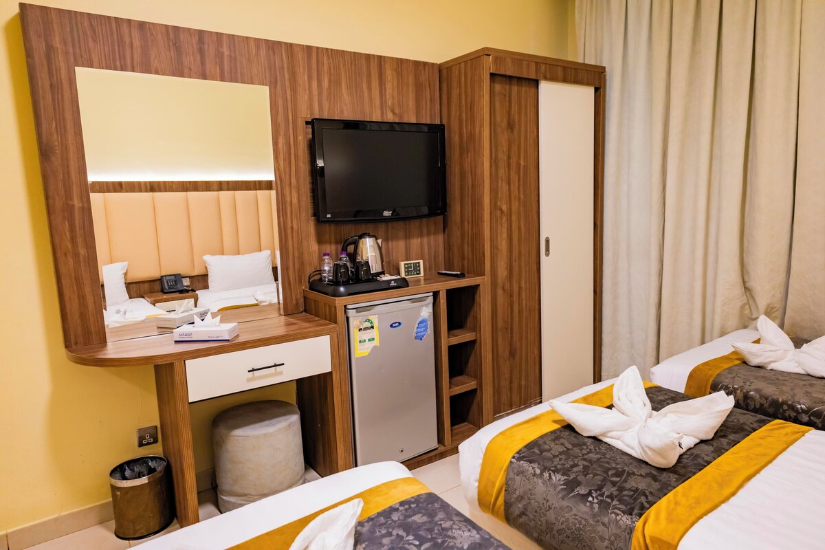 A hotel room with a wooden desk, a television, a mini-fridge, and two beds with towels.