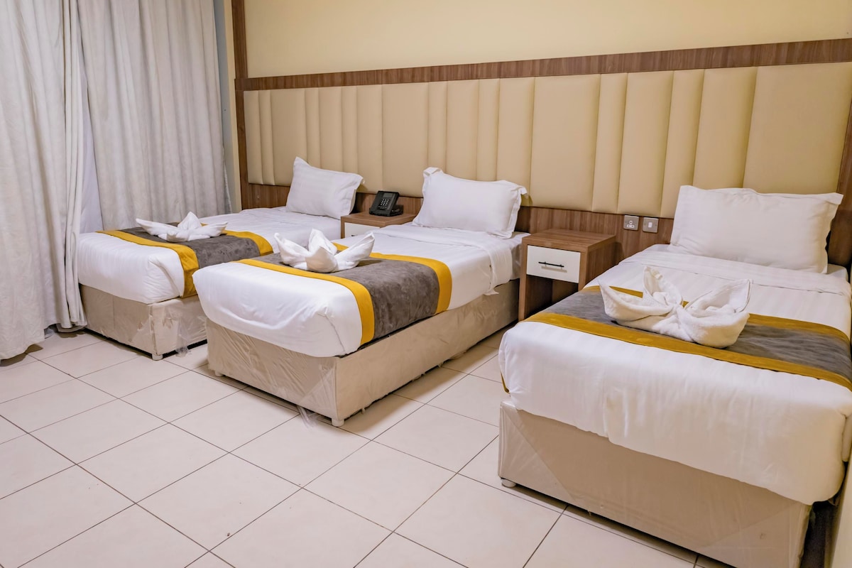 A hotel room with three beds, each with white bedding and a yellow accent. There are white curtains on the windows, and a wooden headboard.
