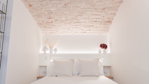 Une chambre avec un lit blanc, des murs blancs, un plafond en briques et une tête de lit blanche.