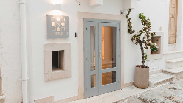 Une entrée d’immeuble avec une porte à double battant grise, un pot de fleurs et une enseigne arborant un logo géométrique.