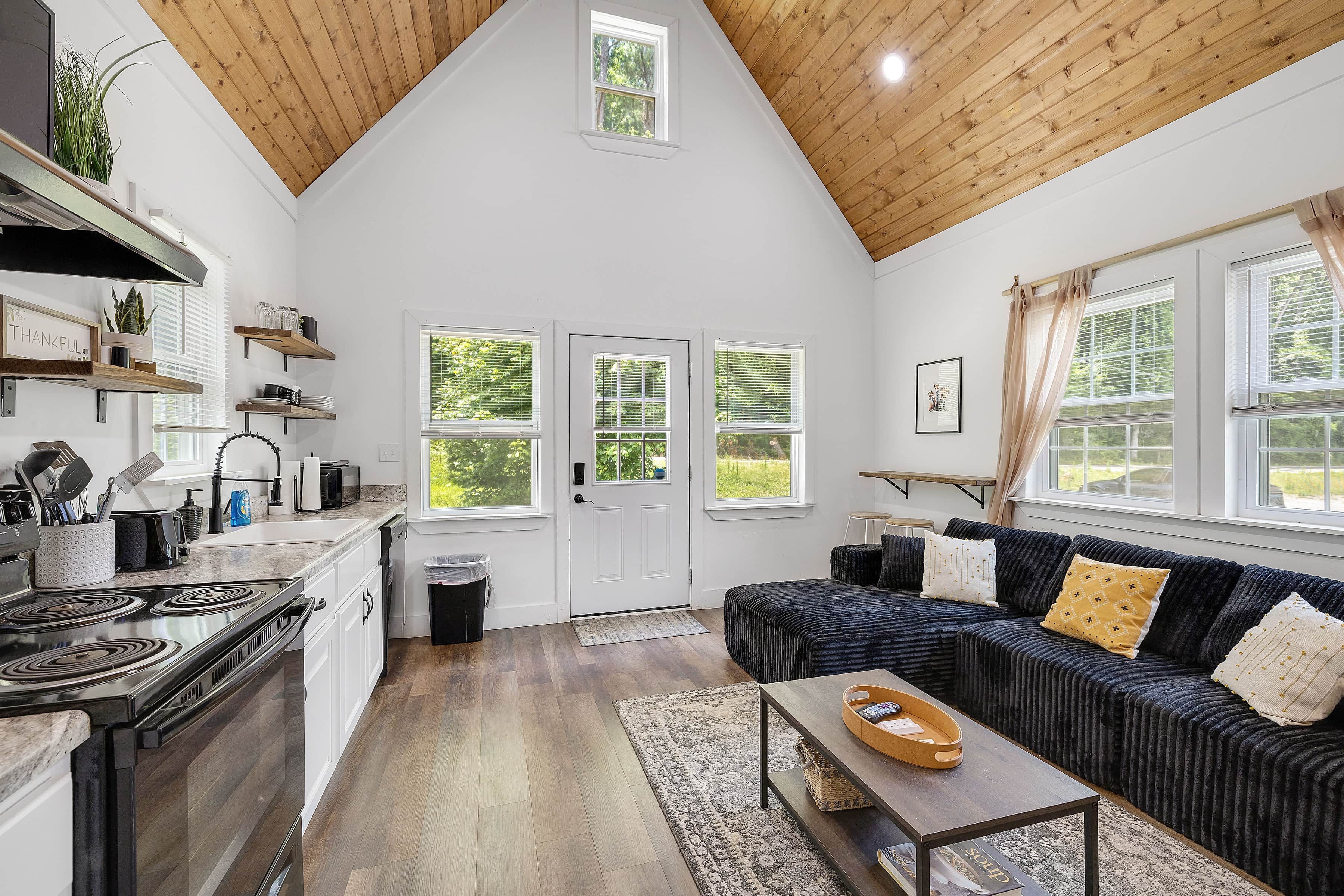 A spacious living area with a high ceiling, a kitchen with modern appliances, and a cozy seating area with a dark-colored sofa and cushions.