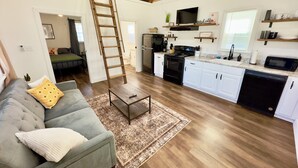 Amplia sala de estar con un entrepiso con cama, una cocina con armarios blancos y techo de madera.