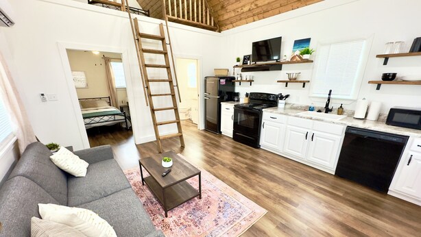 Eine moderne Wohnung mit Schlafgalerie, einer Küche mit weißen Schränken, einem Essbereich und einem Wohnzimmer mit grauem Sofa und Couchtisch.
