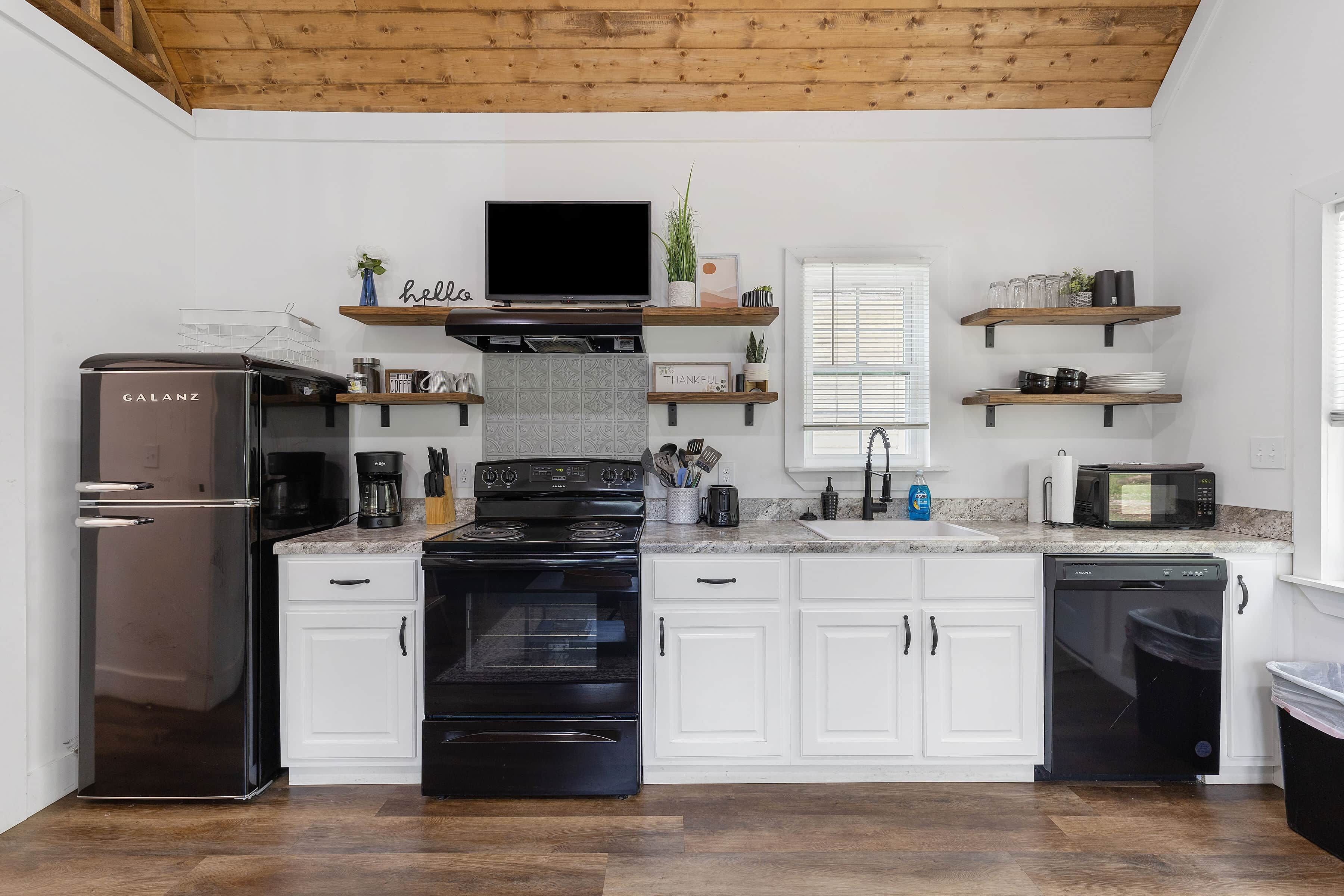 Une cuisine moderne avec des meubles blancs, une cuisinière noire, un réfrigérateur et un four à micro-ondes. On trouve des étagères en bois au-dessus de la cuisinière et une fenêtre avec des stores.