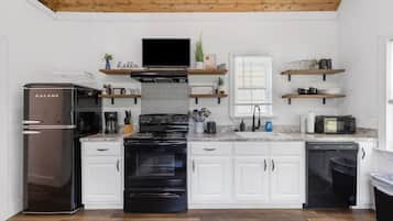 Une cuisine moderne avec des meubles blancs, une cuisinière noire, un réfrigérateur et un four à micro-ondes. On trouve des étagères en bois au-dessus de la cuisinière et une fenêtre avec des stores.