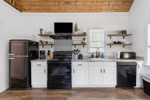 A modern kitchen with white cabinets, a black stove, a refrigerator, and a microwave. There are wooden shelves above the stove and a window with blinds.