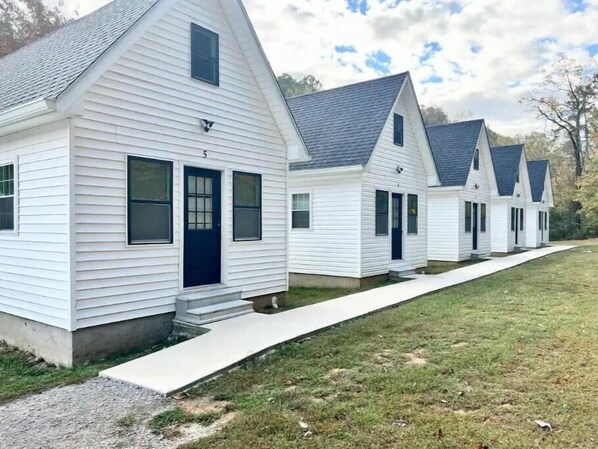 A row of white, single-story houses with dark roofs and blue doors, aligned along a grassy area with a concrete path.