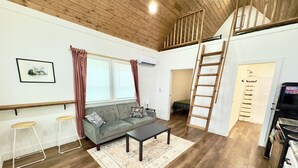 A cozy living room with a sofa, two stools, a coffee table, a wooden ladder, and a staircase leading to an upper level. - Raccoon Cabin: Arcade, Kayaks, Inflatables, & More (Guntersville)