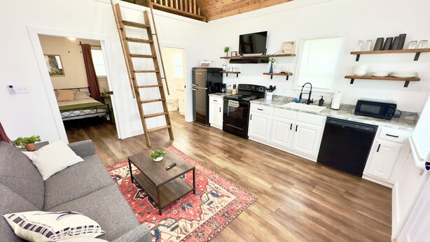 A modern apartment with a loft bedroom, a kitchen with white cabinets, a dining area, and a living room with a gray sofa and a red patterned rug. - Rabbit Cabin: Arcade, Fishing, Kayaks, & More! (Guntersville)