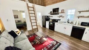 Amplia sala de estar con un entrepiso con dormitorio, piso de madera, cocina con armarios blancos y una alfombra roja estampada.