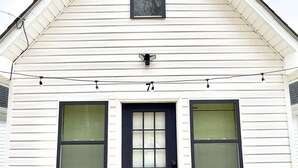 Una casa blanca con una puerta principal oscura, dos ventanas con persianas, un pequeño ventanuera y un accesorio de iluminación visible sobre la puerta.