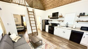 A spacious living area with a gray sofa, a wooden ladder, and a kitchen with white cabinets and a black stove.