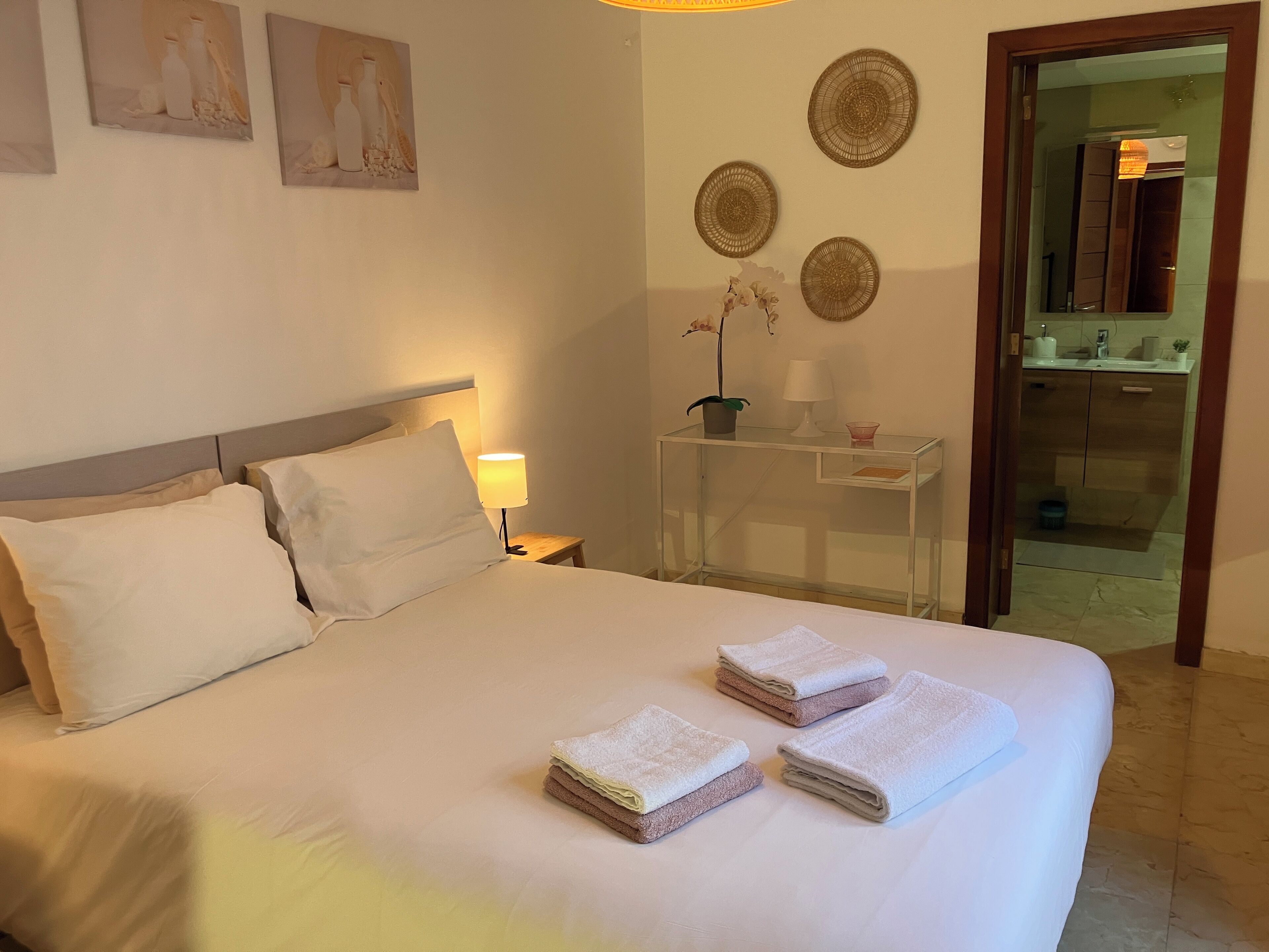 Ein ordentlich bezogenes Bett mit weißen Leinen und zwei gefalteten Handtüchern. Ein Nachttisch mit Lampe und kleiner Topfpflanze ist vorhanden. Der Raum hat ein schlichtes, sauberes Design mit einem Spiegel und Wandbildern über dem Bett.