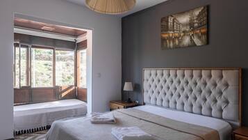 A bedroom with a large bed, a window with a view of greenery, a wall-mounted painting, and a bedside table with a lamp.